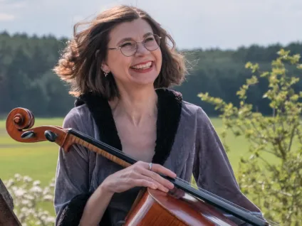 Helke Menter sitter utomhus med en cello i famnen 
